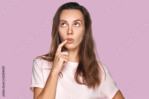 People and thoughts concept. Attractive Caucasian woman looks pensive upwards, keeps forefinger near lips, plans day, has something in mind on lavender studio. Clever woman thinks about new project