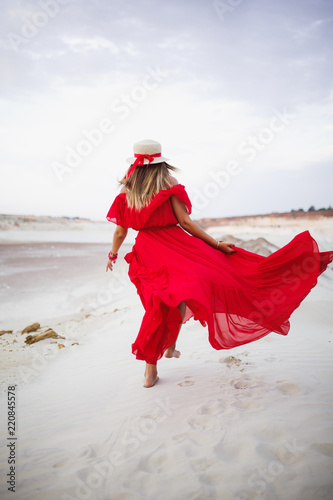 Beautiful young girl in red dress and fedora hat with stripe walking in the desert with white sand