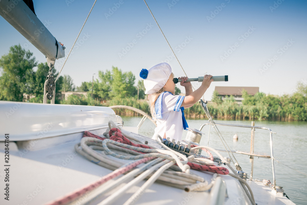 Fototapeta premium Funny little baby captain on board of sailing yacht watching offshore sea on summer cruise. Travel adventure, yachting with child on family vacation. Kid clothing in sailor style, nautical fashion.
