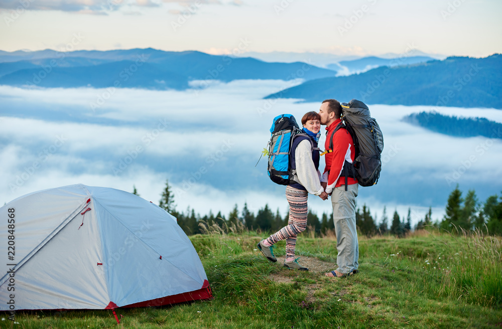 Guy kissing the girl on the blurred background of the mountains in the morning. Couple with backpacks near a tent during active holidays in the mountains