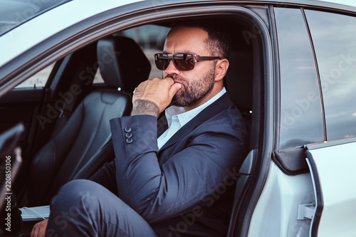 Confident businessman in sunglasses dressed in a formal suit sitting on the front seats in the luxury car. © Fxquadro