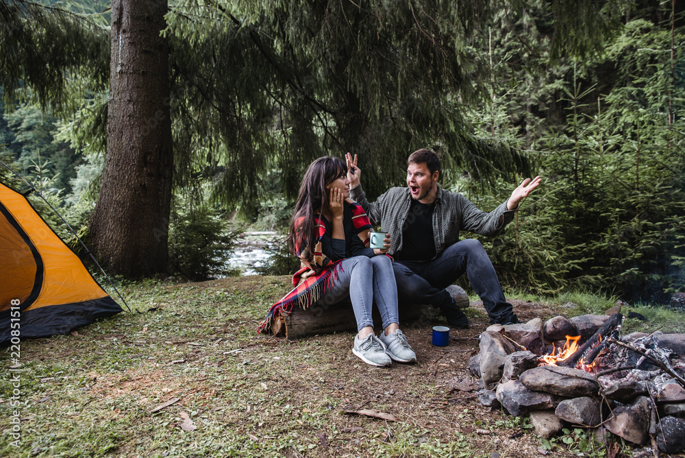 couple sitting near camp fire and drinking tea and telling stories . tent and suv on background couple sitting near camp fire and drinking tea and telling stories . tent and suv on background