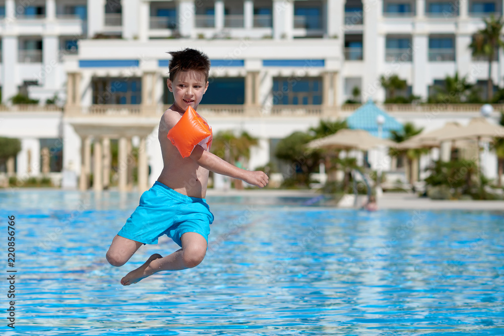 Caucasian boy in floating sleeves jumping into water in swimming pool at resort. He is spinning in air.