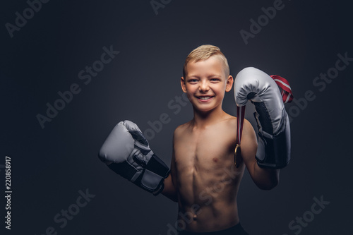 Joyful shirtless boy boxer in boxing gloves holds a golden medal posing in a studio. © Fxquadro