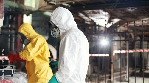 Caucasian guy wearing white protective suit and green rubber gloves finishing work. Two young men in gas masks looking directly at camera. Invisible danger.