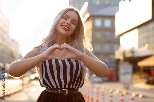 Positive blonde girl with red lips making heart sign with her fingers at the street