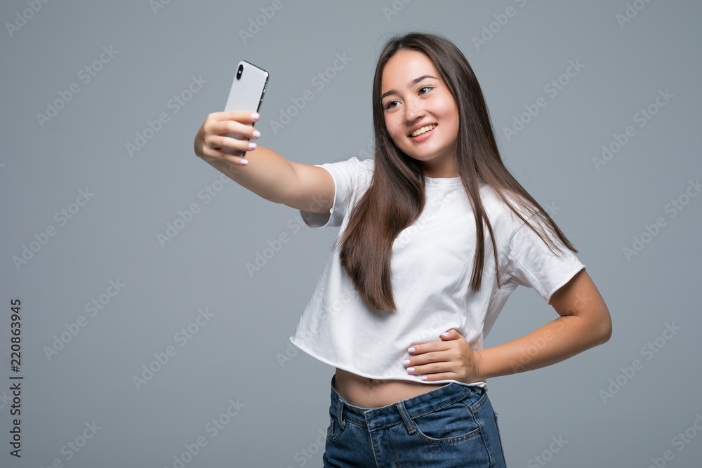Sociable beautiful asian girl taking selfie or speaking on video call using cell phone isolated over gray background