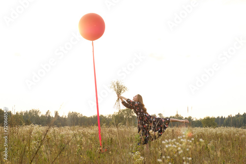 Young girl in a beautiful dress with flowers with a red balloon on a leash in the field at sunset.