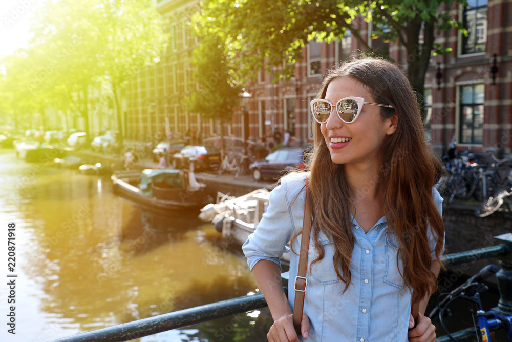 Portrait of beautiful cheerful girl with sunglasses looking to the side on one of typical Amsterdam channels, Netherlands. Light flare vintage filter.