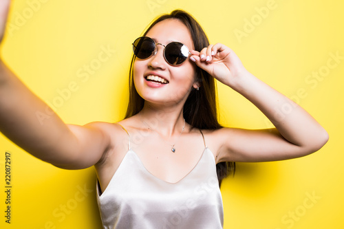 Young cute asian girl making a selfie, standing on the bright yellow background and smiling