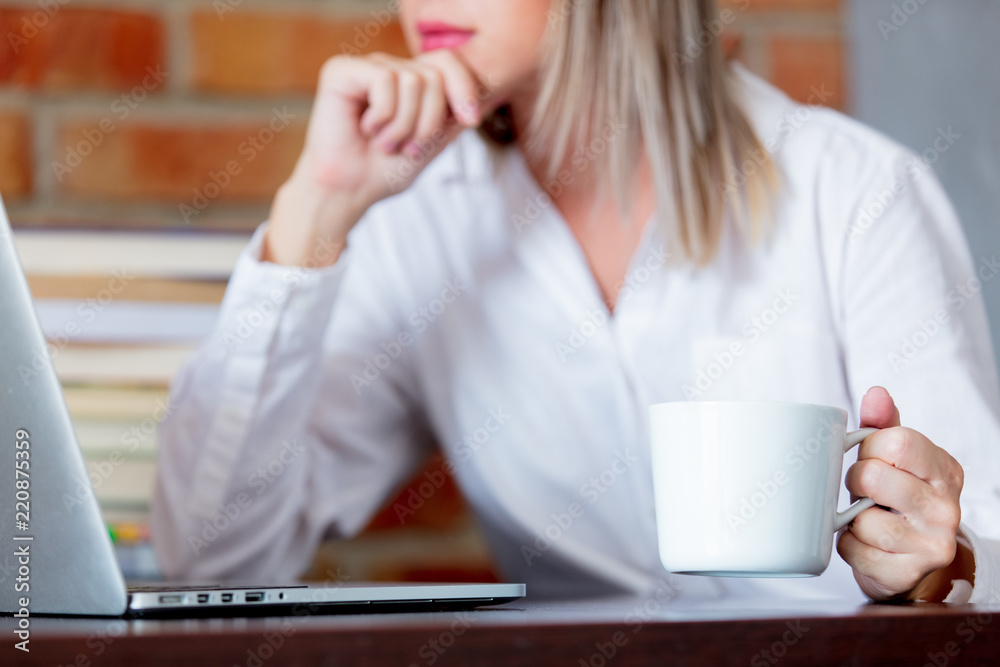 Young businesswoman sitting on working place with laptop computer and books holding a hot drink. Brick wall on background Young businesswoman sitting on working place with laptop computer and books holding a hot drink. Brick wall on background