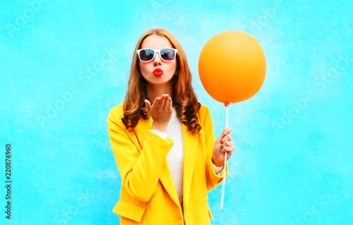 portrait beautiful woman with balloons sends an air kiss in yellow coat on colorful blue background