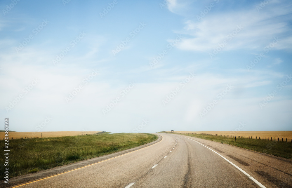 A curving highway between fenced pasture land under a blue wispy sky in a summertime Montana landscape