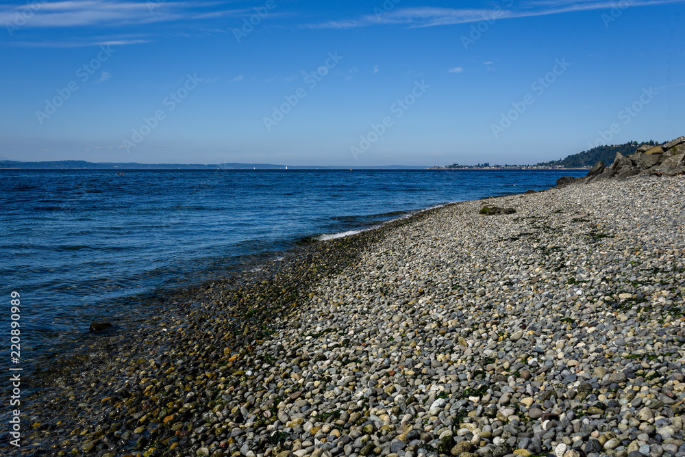 Rocky Puget Sound beach landscape, small rocks, calm sea with small waves, distant island and blue sky with clouds
