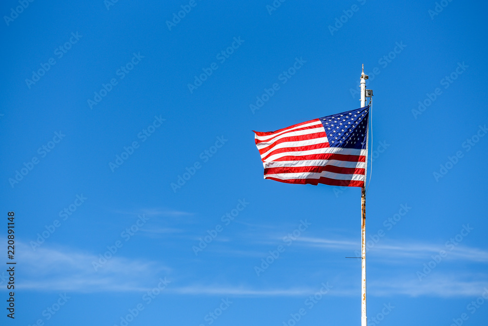 American flag on top white flagpole, flying in the breeze, blue sky background, wispy white cloud, stars and stripes, red, white, and blue

