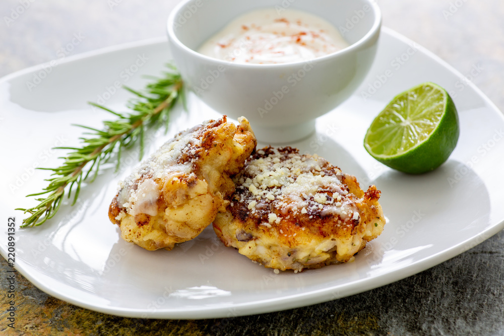 Two shrimp cakes on a white plate with a lemon garlic dipping sauces lemon wedge and a stem or rosemary on a wooden table. Two shrimp cakes on a white plate with a lemon garlic dipping sauces lemon wedge and a stem or rosemary on a wooden table.