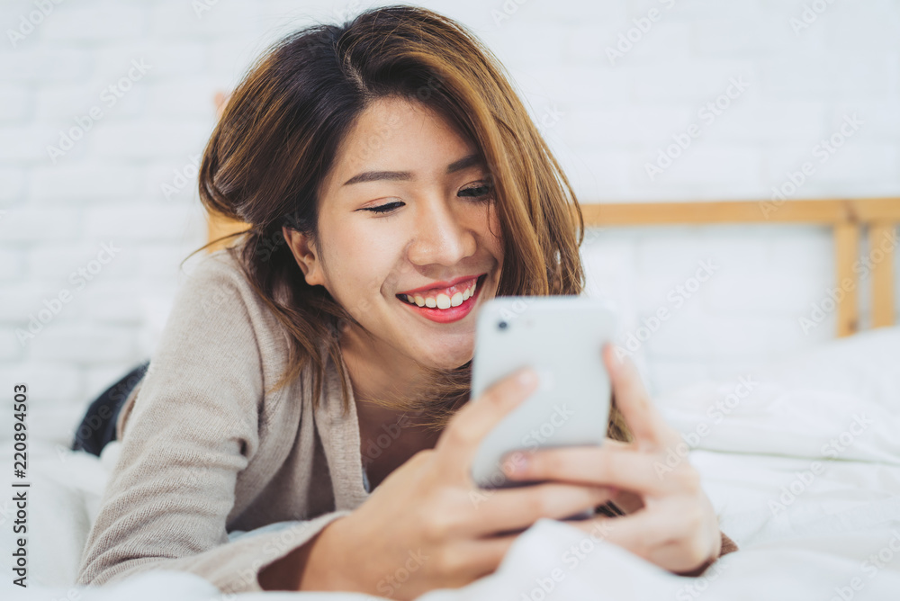 Happy Asian women are using smart phone on the bed in morning. Asian woman in bed checking social apps with smartphone. Smiling woman surfing net with cellphone at home. Mobile addict concept.