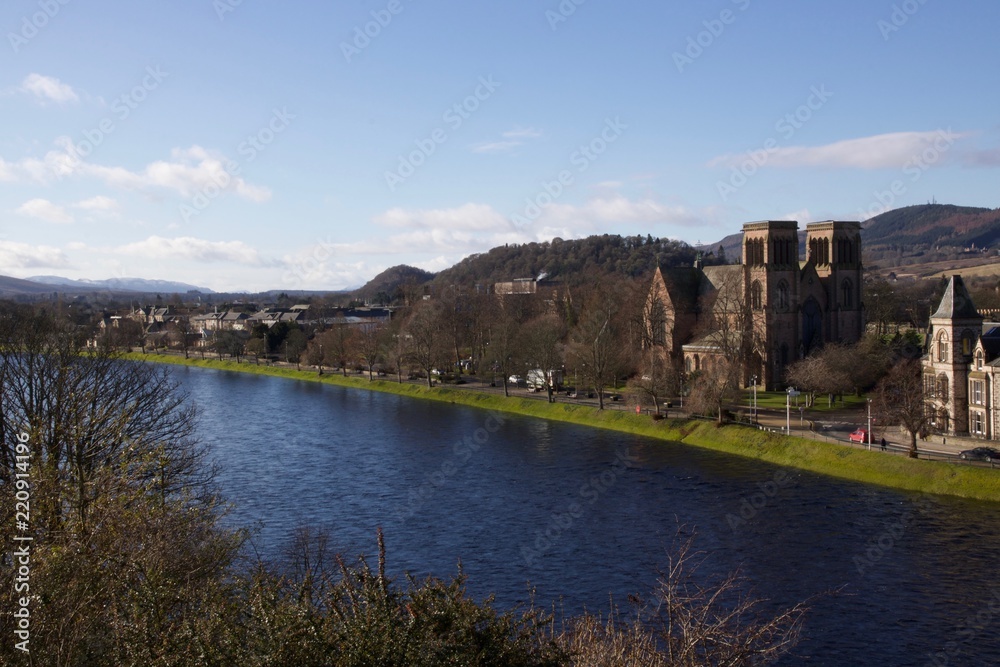 A cathedral in Inverness runs along the Scottish city river on a sunny day with blue skies and green trees in the foreground