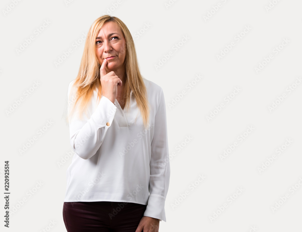 Middle age blonde woman with white shirt having doubts and with confuse face expression while looking up. Questioning an idea on isolated grey background Middle age blonde woman with white shirt having doubts and with confuse face expression while looking up. Questioning an idea on isolated grey background