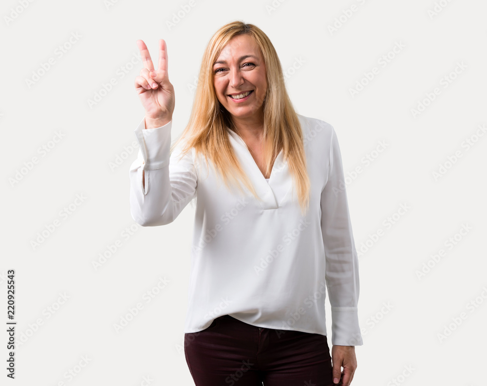 Middle age blonde woman with white shirt smiling and showing victory sign with a cheerful face on isolated grey background Middle age blonde woman with white shirt smiling and showing victory sign with a cheerful face on isolated grey background