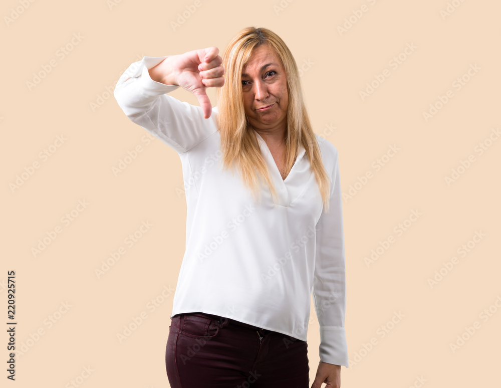 Middle age blonde woman with white shirt showing thumb down sign with negative expression. Sad expression on isolated ocher background Middle age blonde woman with white shirt showing thumb down sign with negative expression. Sad expression on isolated ocher background