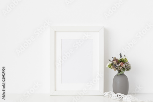 Fototapeta Naklejka Na Ścianę i Meble -  White a5 portrait frame mockup with dried field wild flowers in vase and vintage lace on white wall background. Empty frame, poster mock up for presentation design. 