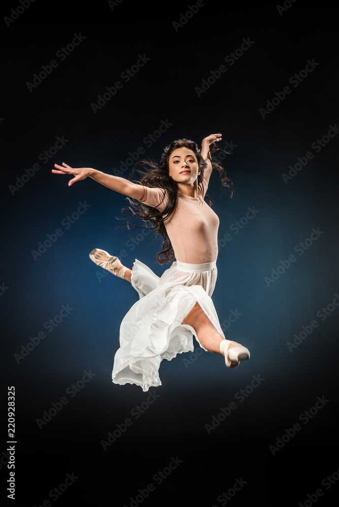 young ballerina in elegant clothing jumping on dark backdrop
