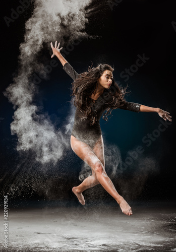 beautiful ballerina in black bodysuit jumping on dark background with talc powder around
