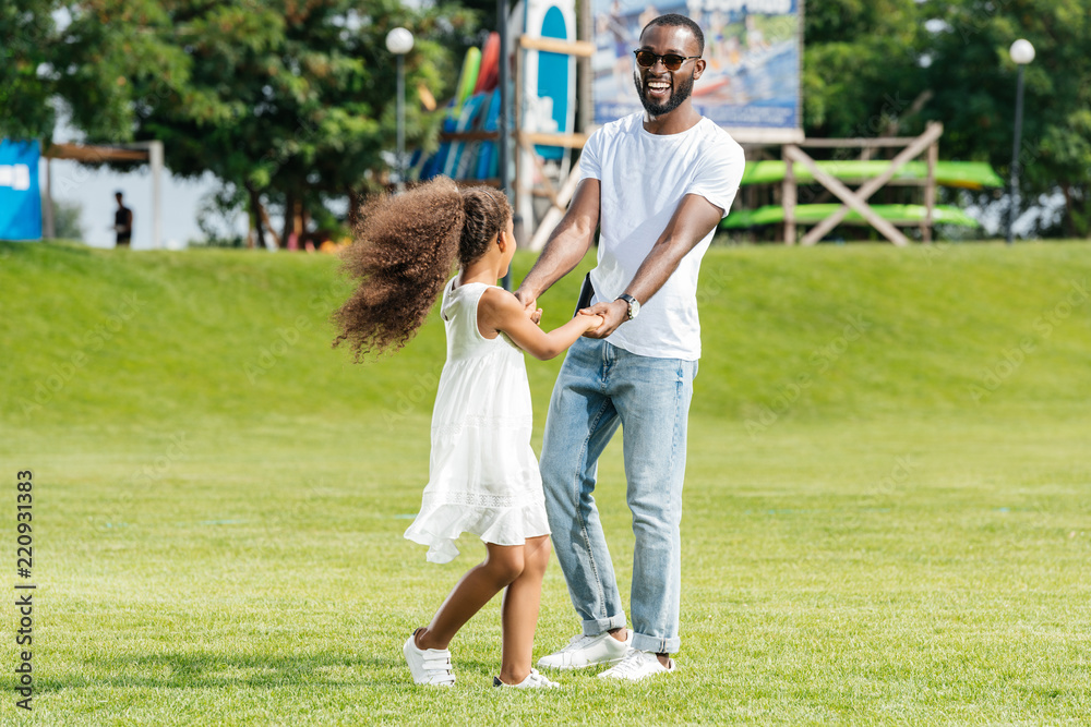 Fototapeta premium happy african american father and daughter holding hands and spinning around in amusement park