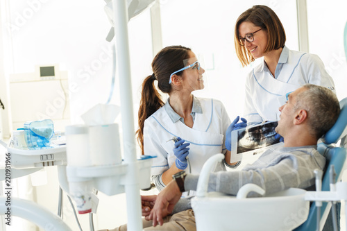 Dentist and his assistant in dental office talking with female patient and preparing for treatment.Examining x-ray image.