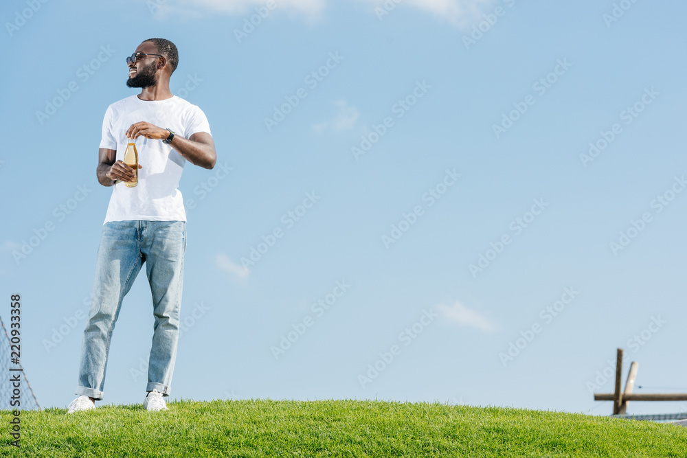 smiling handsome african american man opening soda bottle on green hill and looking away