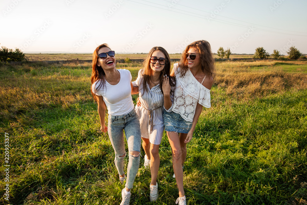 Three gorgeous young women in sunglasses dressed in the beautiful clothes stand in the field and smiling on a sunny day. Three gorgeous young women in sunglasses dressed in the beautiful clothes stand in the field and smiling on a sunny day.
