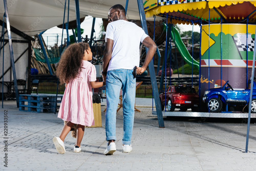 back view of african american police officer walking with daughter and holding police badge at amusement park