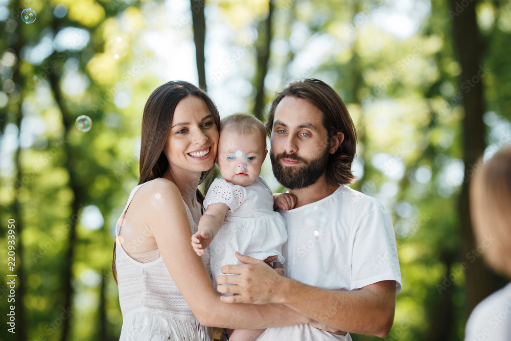 Easygoing young parents dressed in the white clothes are holding daughter in the arms and smiling outdoor. Easygoing young parents dressed in the white clothes are holding daughter in the arms and smiling outdoor.