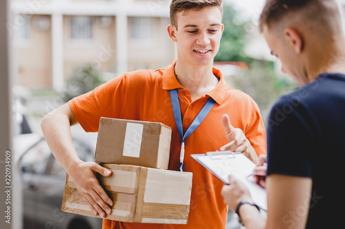 A smiling person wearing an orange T-shirt and a name tag is delivering a parcel to a client, who is putting his signature on the receipt. Friendly worker, high quality delivery service.