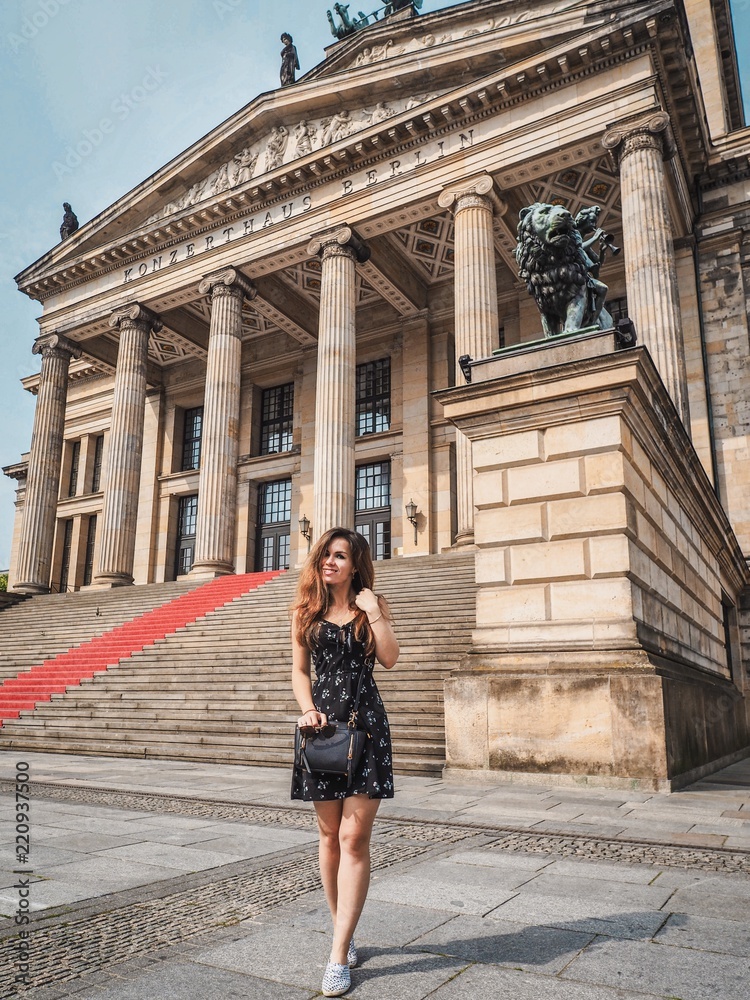 A girl in a black dress, with a small bag and sneakers, with long hair stands on the background of a Museum with columns, a red carpet and statues in Berlin