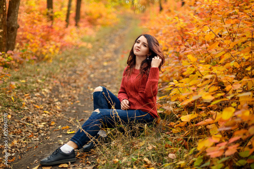 young beautiful woman in red knitted sweather sitting on ground in autumn park with yellow and red leaves