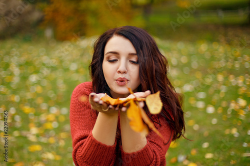 Portait of young beautiful woman in red knitted sweather laying on grass and blows leaves on camera from her hands at autumn park with yellow leaves
