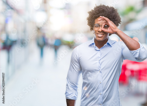 Afro american business man wearing glasses over isolated background doing ok gesture with hand smiling, eye looking through fingers with happy face.