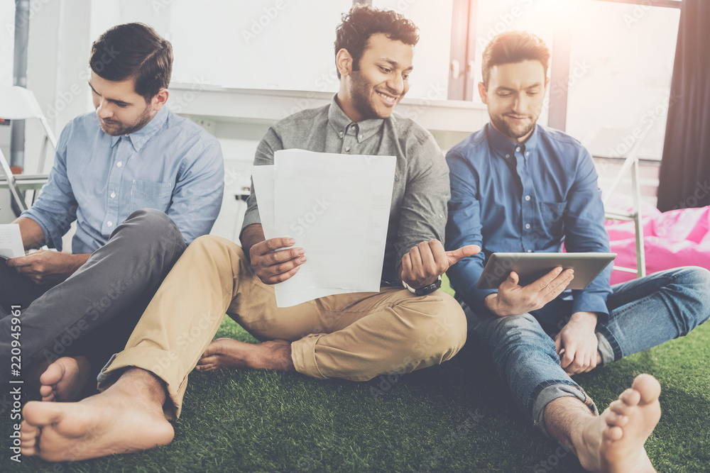 Smiling young barefoot businessmen sitting with papers and using digital tablet, business teamwork concept Smiling young barefoot businessmen sitting with papers and using digital tablet, business teamwork concept