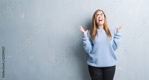 Young adult woman over grey grunge wall wearing glasses crazy and mad shouting and yelling with aggressive expression and arms raised. Frustration concept.