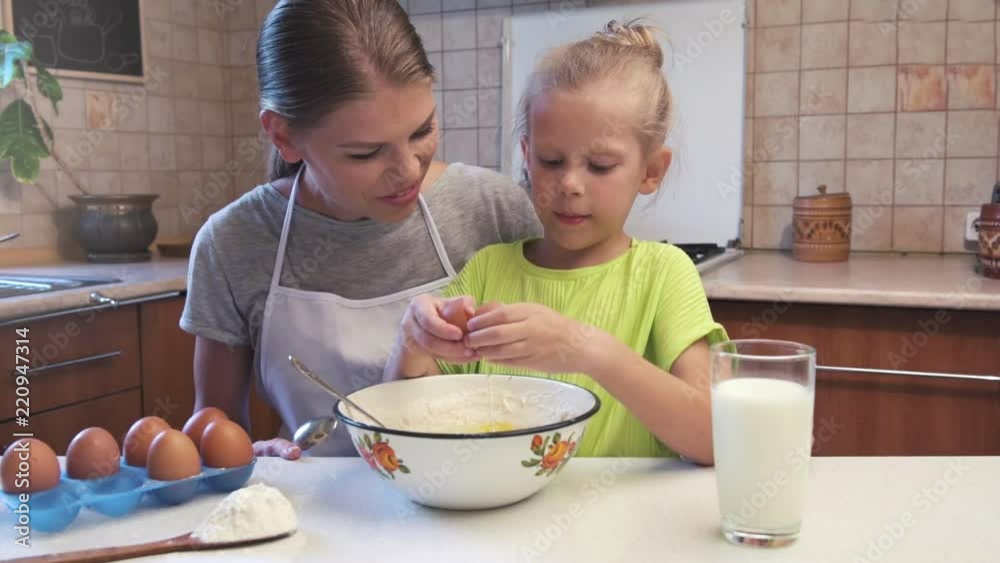 Little daughter breaking egg into dough, helping mother preparing pie at the table. Little daughter breaking egg into dough, helping mother preparing pie at the table.