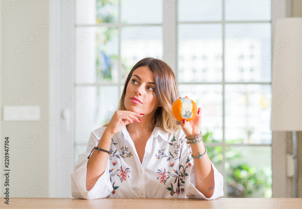 Young woman at home holding rotten orange serious face thinking about question, very confused idea Young woman at home holding rotten orange serious face thinking about question, very confused idea