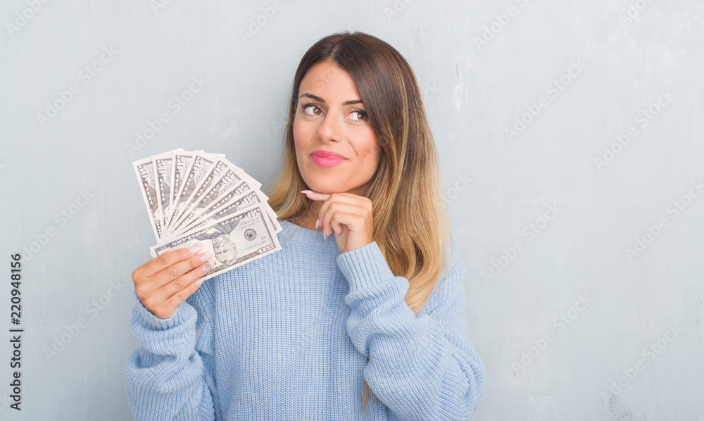 Young adult woman over grey grunge wall holding dollars serious face thinking about question, very confused idea Young adult woman over grey grunge wall holding dollars serious face thinking about question, very confused idea