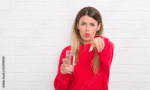 Young adult woman over white brick wall drinking glass of water pointing with finger to the camera and to you, hand sign, positive and confident gesture from the front