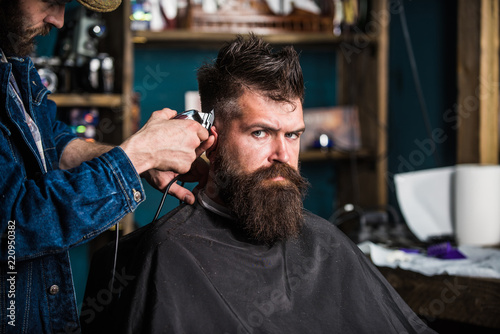 Stylish haircut concept. Hands of barber with clipper close up. Hipster bearded client on strict face getting hairstyle. Barbers hand with hair clipper trimming. Client with beard on salon background