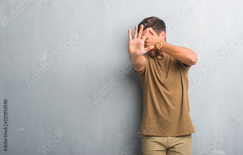 Handsome young man over grey grunge wall covering eyes with hands and doing stop gesture with sad and fear expression. Embarrassed and negative concept.