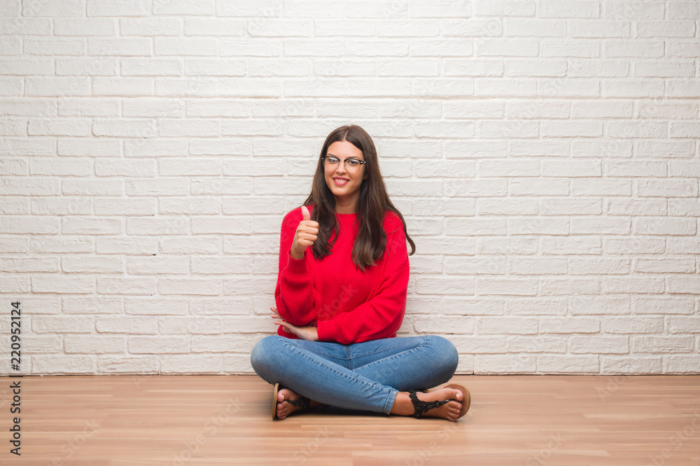 Young brunette woman sitting on the floor over white brick wall happy with big smile doing ok sign, thumb up with fingers, excellent sign Young brunette woman sitting on the floor over white brick wall happy with big smile doing ok sign, thumb up with fingers, excellent sign
