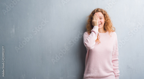 Young redhead woman over grey grunge wall wearing pink sweater covering one eye with hand with confident smile on face and surprise emotion. © Krakenimages.com