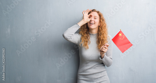 Young redhead woman over grey grunge wall holding flag of China with happy face smiling doing ok sign with hand on eye looking through fingers